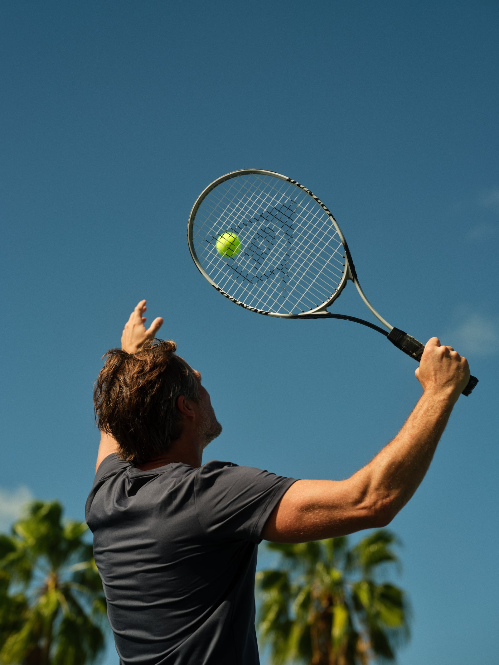 A man perfecting his serve under the clear blue sky at Grand Reserve's luxurious BATÚ Sports Center, surrounded by lush tropical greenery