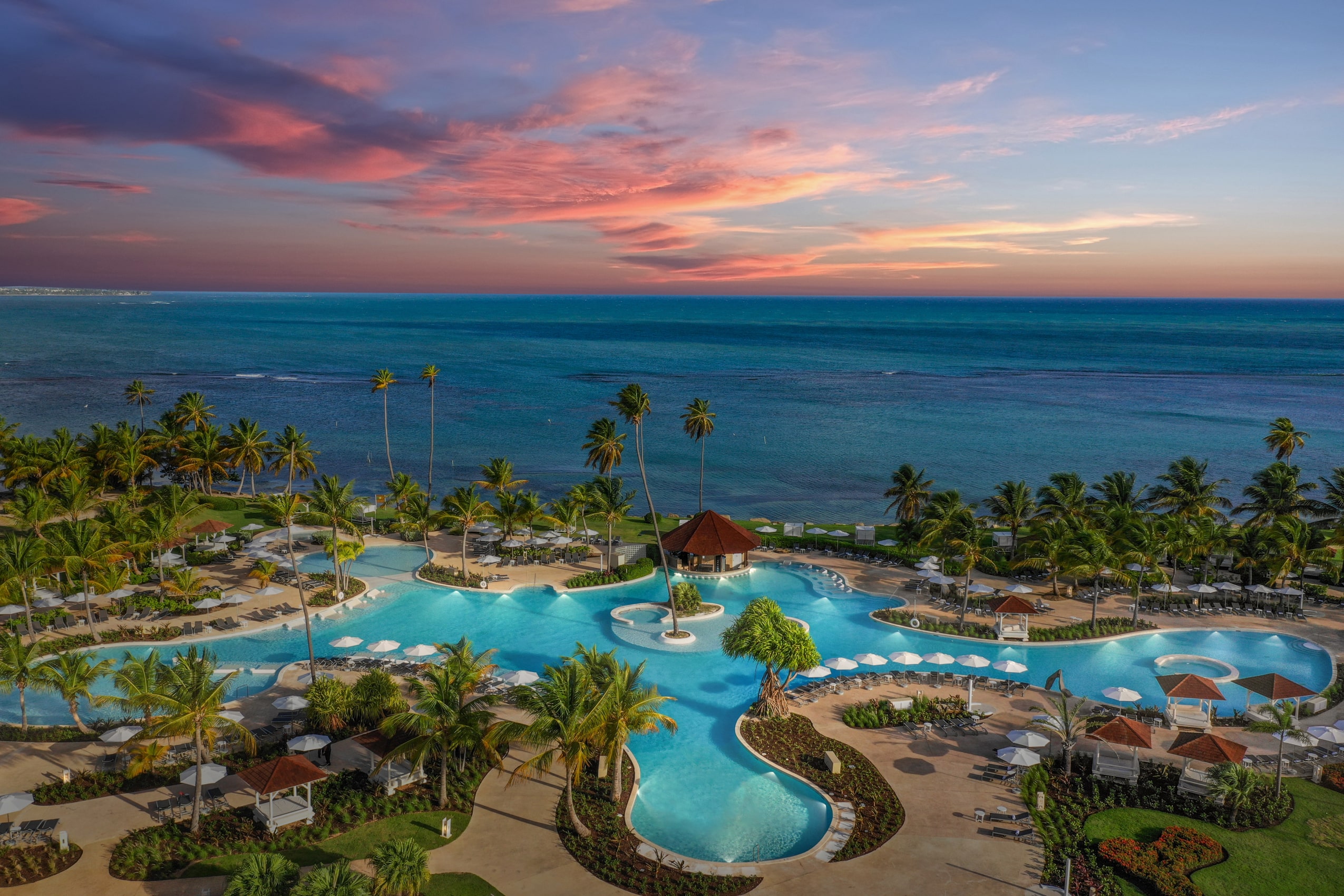 A breathtaking aerial view of Grand Reserve at dusk, showcasing the resort's expansive pool complex with swaying palm trees and the serene Caribbean Sea in the background, under a sky painted with vibrant hues of sunset