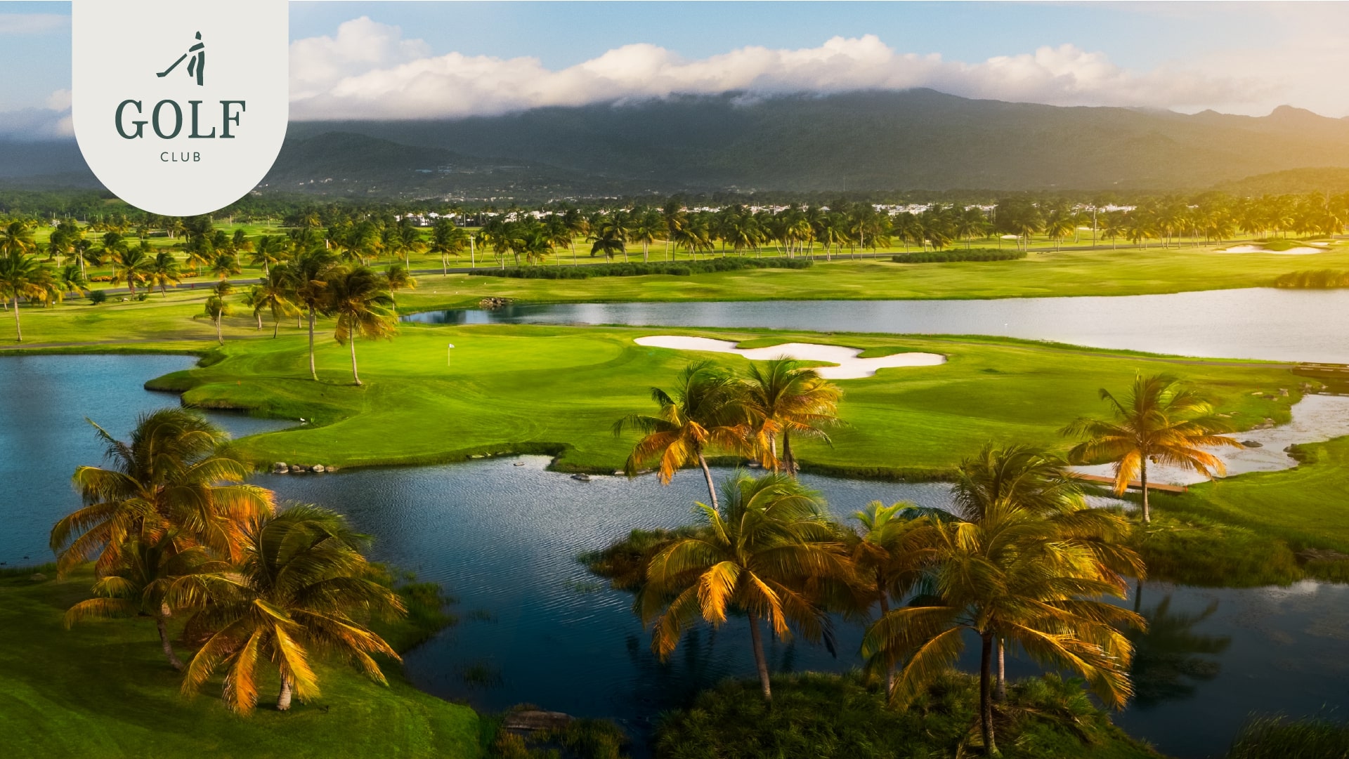 Aerial view of a lush green golf course with water features at Grand Reserve, surrounded by palm trees and mountains in the distance.